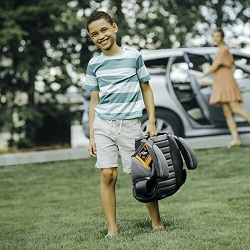 Smiling boy carrying a booster seat on grass with a car in the background.